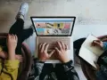 Zespół marketingu realizuje zadania: ktoś pisze na laptopie, ktoś notuje w zeszycie, tworząc kreatywną kampanię.