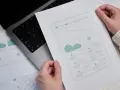 Ręce trzymają szkic strony docelowej (landing page) z elementami interfejsu. Obok leży laptop.