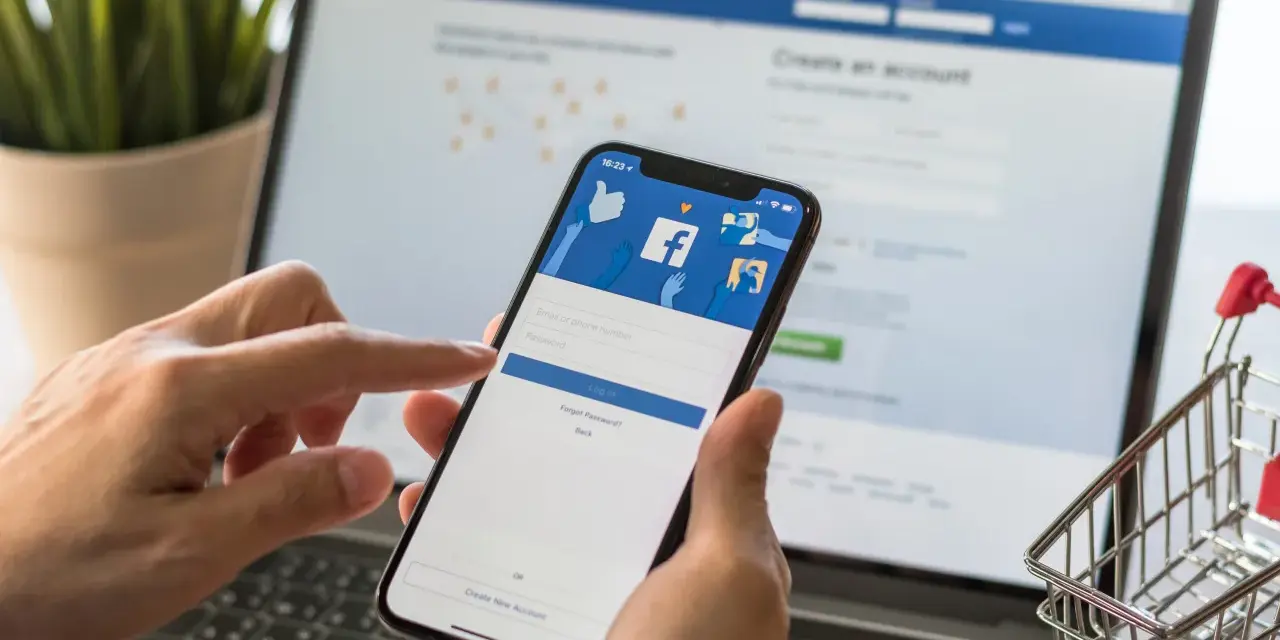 Ręce trzymają telefon z ekranem logowania Facebooka, obok laptopa. Zastanawiasz się, dlaczego strona na Facebooku jest niewidoczna?