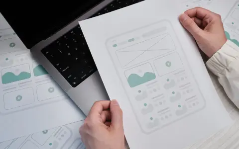 Ręce trzymają szkic strony docelowej (landing page) z elementami interfejsu. Obok leży laptop.