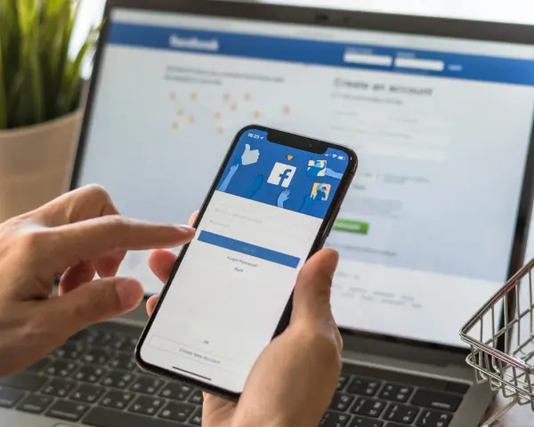 Ręce trzymają telefon z ekranem logowania Facebooka, obok laptopa. Zastanawiasz się, dlaczego strona na Facebooku jest niewidoczna?