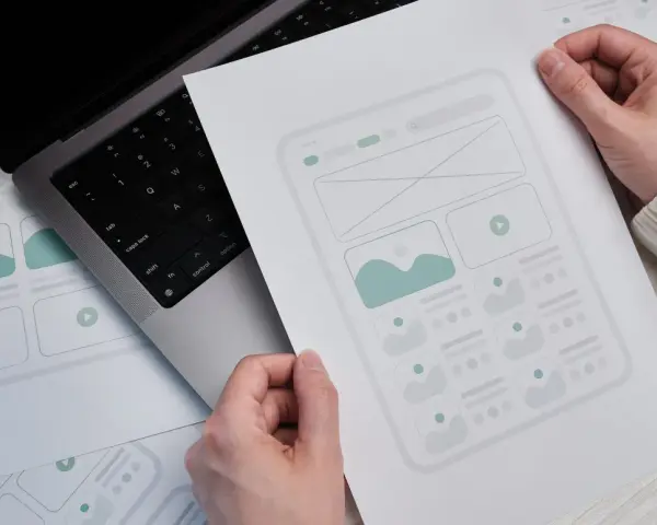 Ręce trzymają szkic strony docelowej (landing page) z elementami interfejsu. Obok leży laptop.