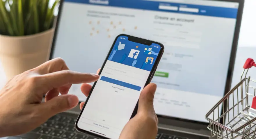Ręce trzymają telefon z ekranem logowania Facebooka, obok laptopa. Zastanawiasz się, dlaczego strona na Facebooku jest niewidoczna?