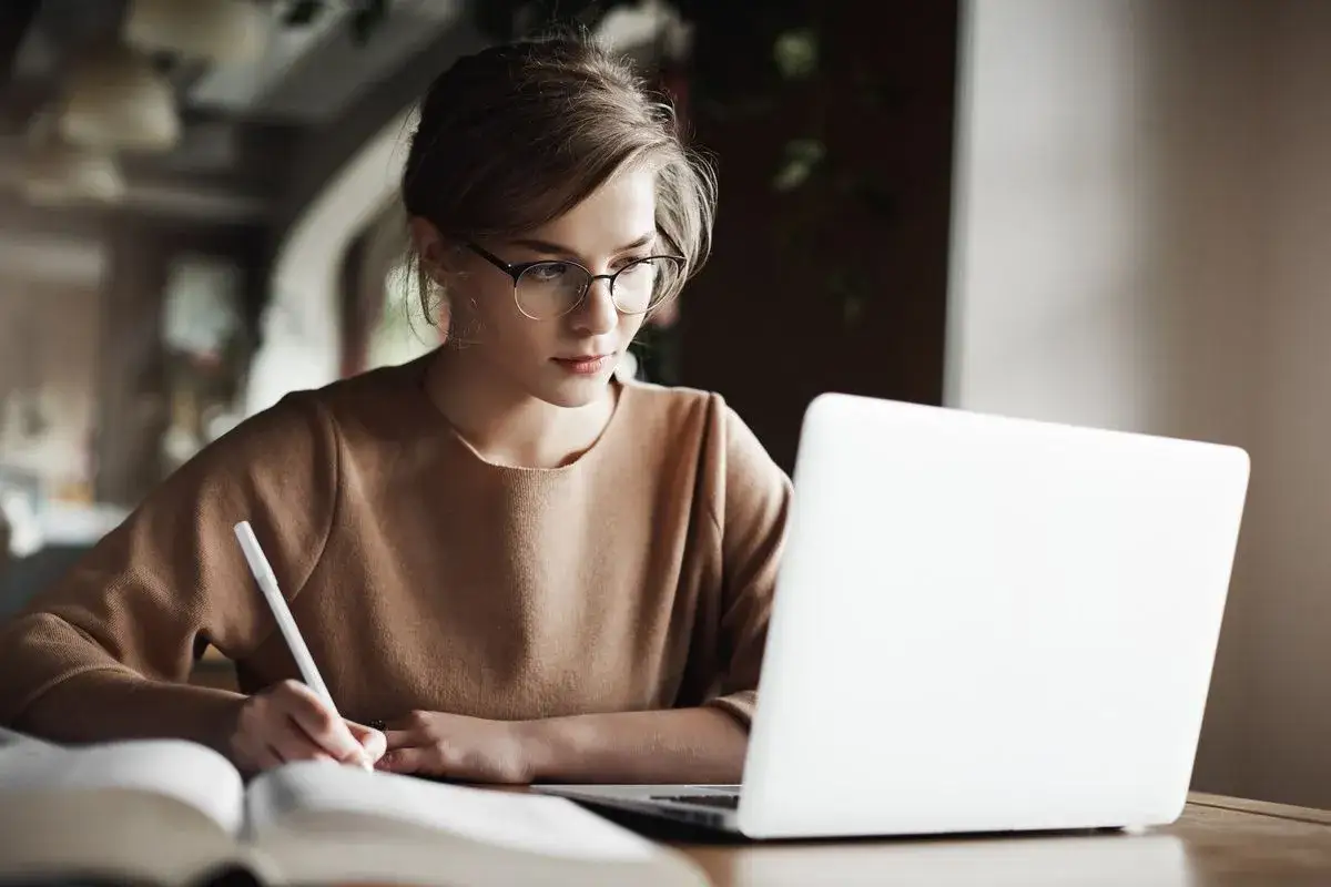 Młoda kobieta w okularach pisze w zeszycie, pracując przy laptopie. Czy tak wygląda bloger kto to?